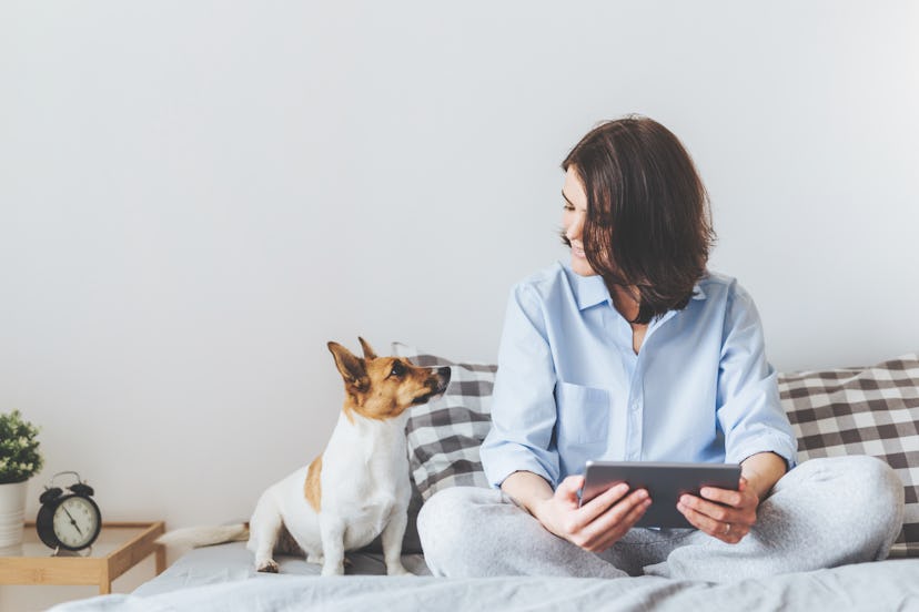 A woman sitting on a bed and holding a tablet and a dog sitting next to her and looking at her
