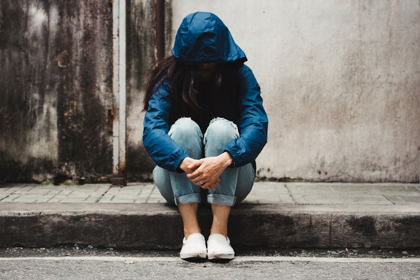 A woman dealing with depression wearing a blue hoodie covering her face while sitting on a sidewalk.