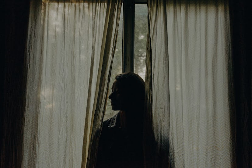 A woman struggling with social anxiety in a dark room, standing in front of a slightly open curtain
