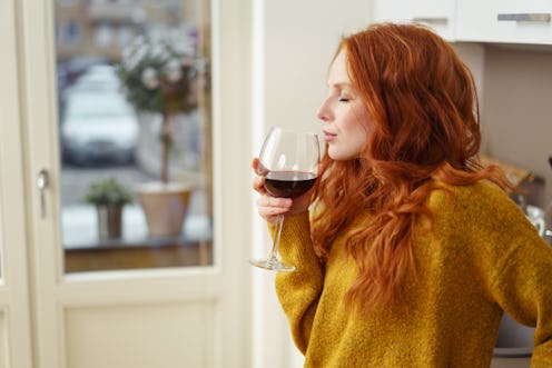 A redheaded woman drinking a glass of red wine while standing. 