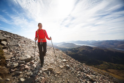 A woman wearing a hiking outfit walking by supporting herself with trekking poles on a rocky mountai...