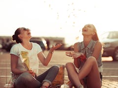 Two best friends smile while they sit on the ground and toss chips in the air at sunset.