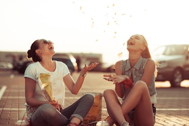 Two best friends smile while they sit on the ground and toss chips in the air at sunset.