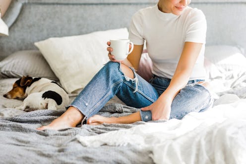 A woman wearing jeans and a white shirt sitting on a bed while drinking coffee before a nap