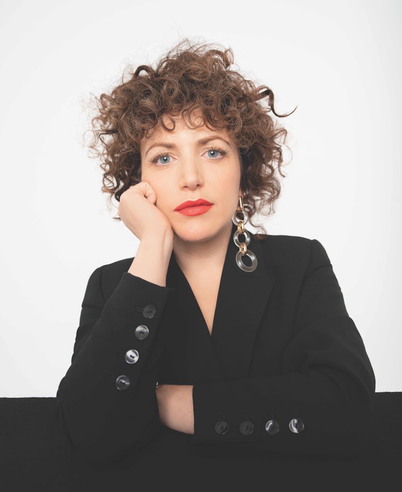 Annie mac in a black blazer sitting at a table with her head resting on her hand in front of a white...