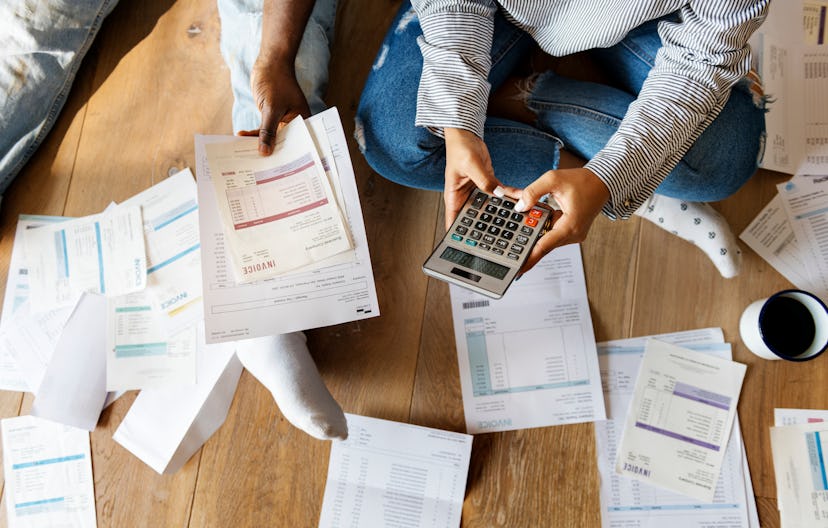 A man and a woman sitting on a floor with lots of papers and bills around them tryin to pay off thei...