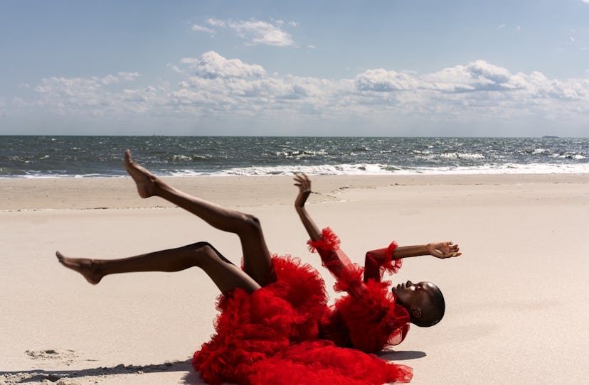 A model lying with her legs and arms lifted in a red Valentino dress with tulle and frills on a beac...
