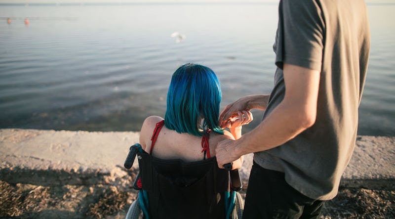 A woman with disabilities sitting in a wheelchair next to a man holding her hand on a shore.