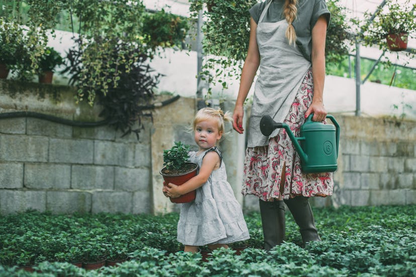 A mom and a daughter in a garden; the mother is carrying a watering can, and the girl is carrying a ...