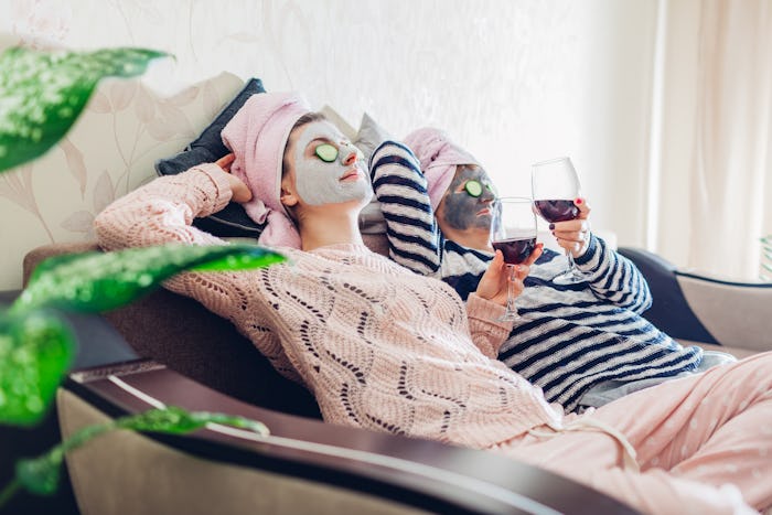 A mother and her daughter relax on a couch while holding wine glasses and wearing face masks.