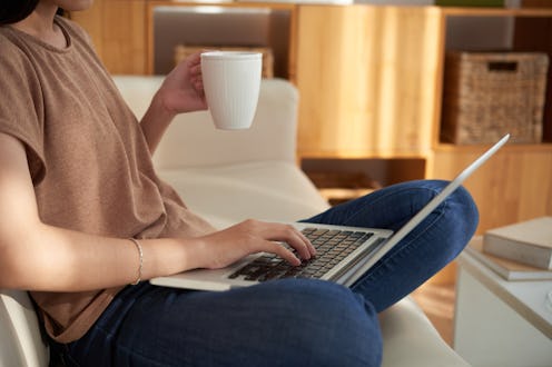 A woman holding a white cup and a laptop in her lap while sitting on a couch