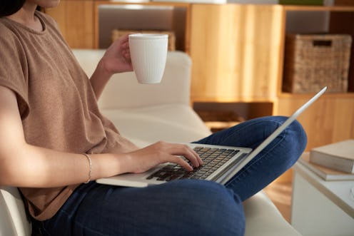 A woman holding a white cup and a laptop in her lap while sitting on a couch