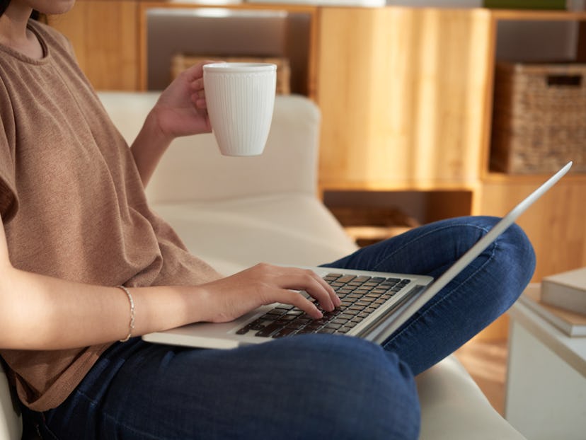 A woman holding a white cup and a laptop in her lap while sitting on a couch