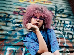 Young woman with pink hair in front of a graffitied background slaying the millennial game on social...