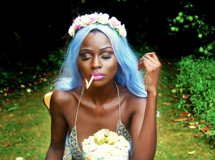 Michelle O Faith with blue hair wearing a floral tiara and a summer dress sitting in a forest smokin...