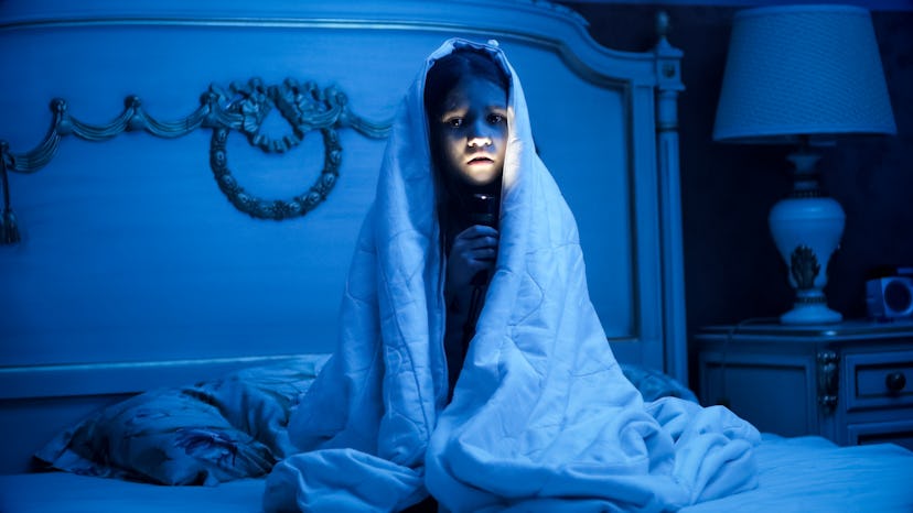A woman sitting on a bed with a blanket over her head and putting light on her face