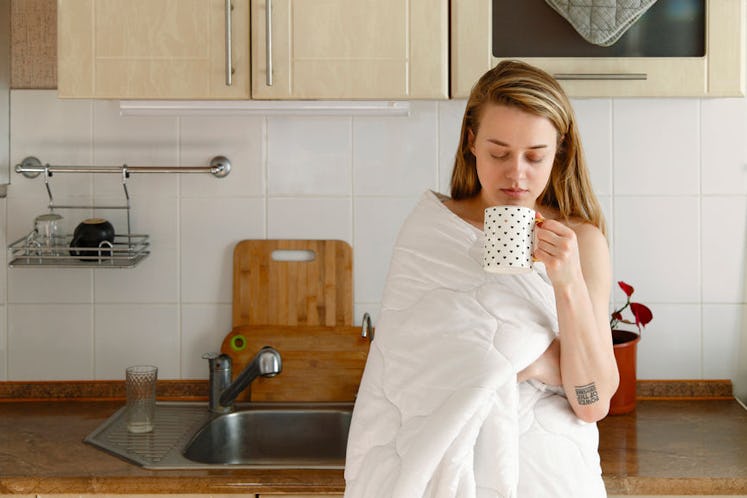 A girl leaning on the kitchen counter wrapped in a blanket while holding a cup of tea. 
