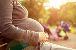 A pregnant woman sitting on a park bench holding her stomach while children play in the background