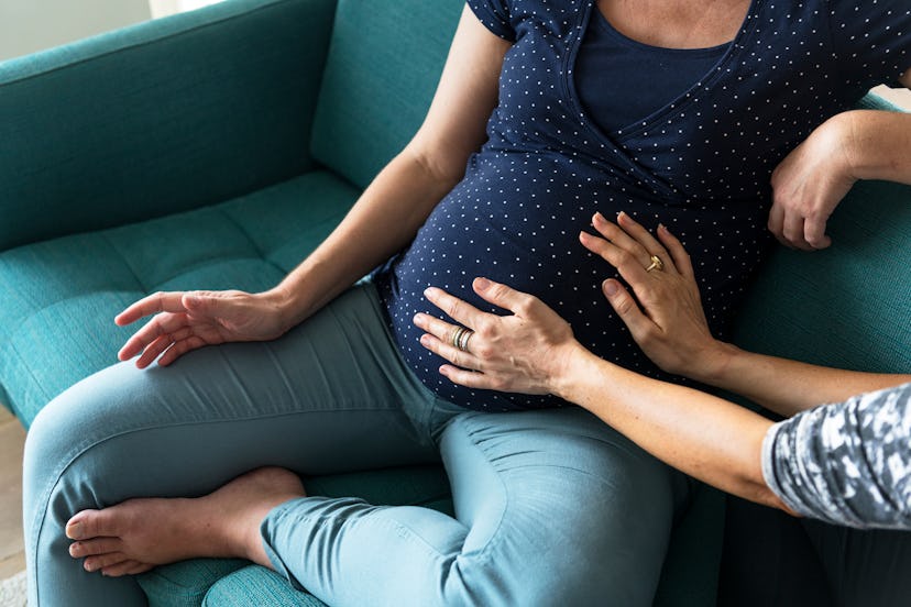 A pregnant woman in a polka dot shirt sitting on the couch while another woman feels her stomach wit...