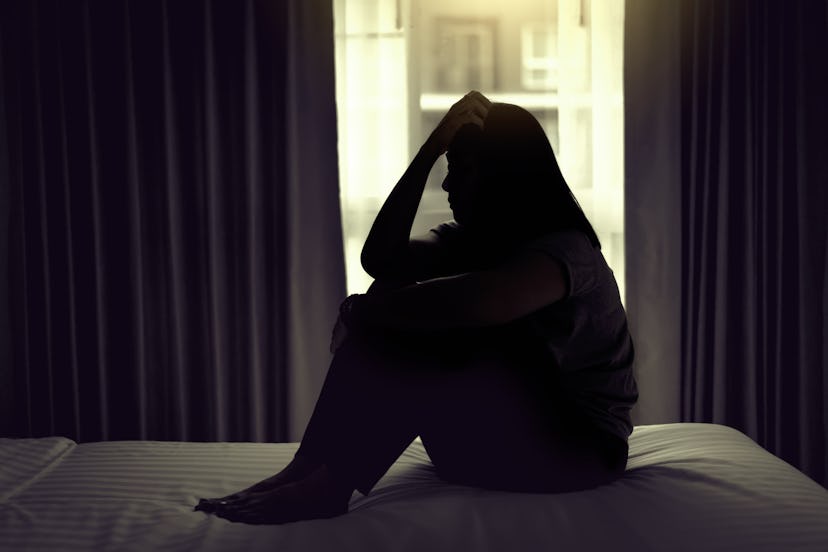 A contre-jour picture of a woman in a silhouette sitting on the bed leaned on her knees 