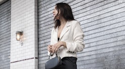 A woman in a white blazer holds a black handbag while standing in front of a grey brick wall