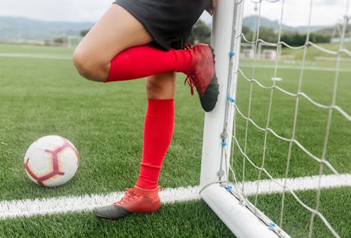 A female football player leaning on a goal while wearing red women's football boots