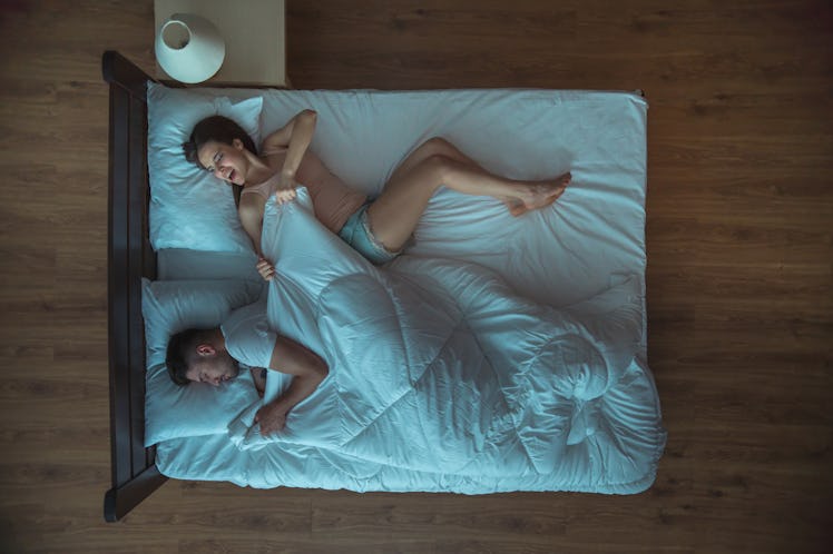 A man pulling the sheets over to his side of the bed while a woman is looking at him confused. 