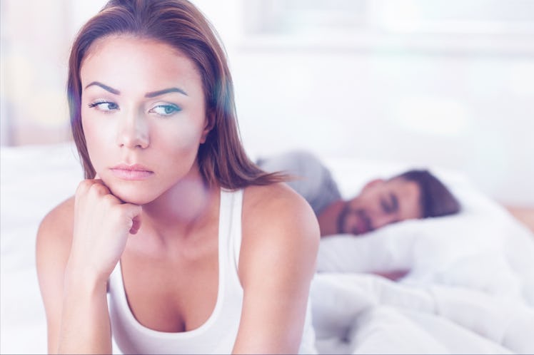 A worried-looking woman sitting on the side of the bed while her boyfriend is peacefully sleeping.