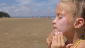 A girl whose mom is terrified of skin cancer, applying sunscreen on her face on a beach