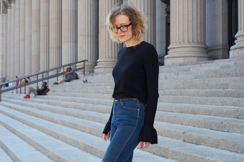 A woman with a blonde curly hair and glasses, wearing a black sweater and blue jeans walking down th...