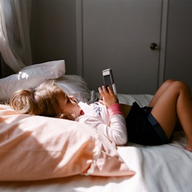 A girl lying in bed while using her phone, sunlight is beaming down on her through the window