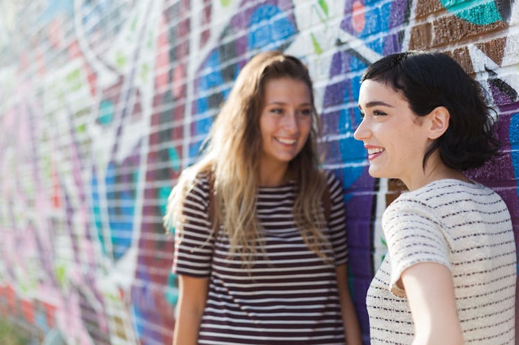 Two women smiling at each other in front of a colorful mosaic wall, wearing casual striped tops.