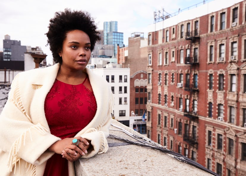 A curly-haired woman in a red shirt and cream coat standing on a rooftop and looking into the distan...