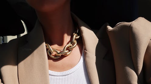 A close-up of a woman's neck, wearing a golden thick chain necklace, a white top and beige blazer