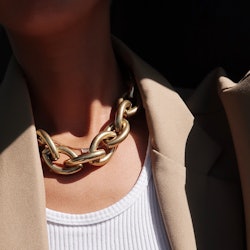 A close-up of a woman's neck, wearing a golden thick chain necklace, a white top and beige blazer