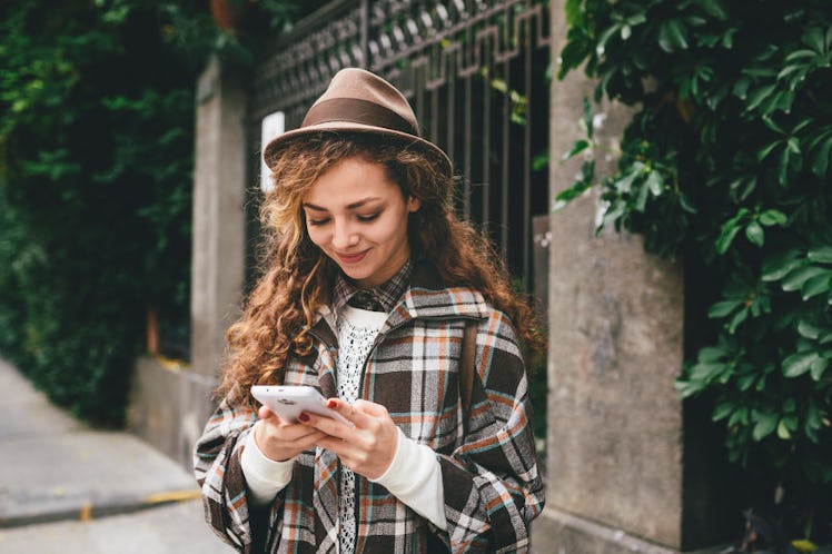 A young woman in a plaid jacket and hat smiles while using her smartphone on a city street, with gre...