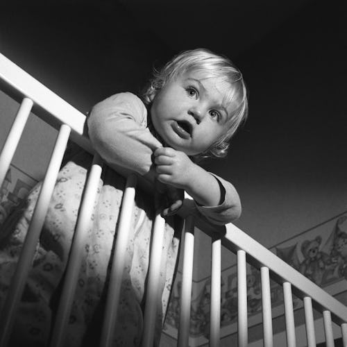A toddler standing in their crib with their hands over the bars trying to get out.