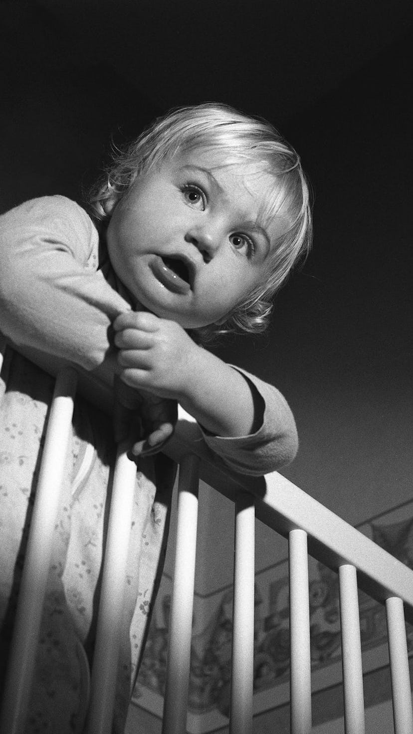 A toddler standing in their crib with their hands over the bars trying to get out.