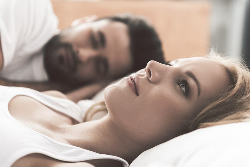 A woman and man lay in bed. The woman is looking up at the ceiling while the man sleeps. 