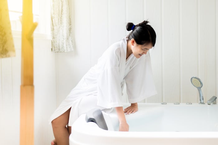 woman in white robe leaning against white tub feeling the water temperature, in a story answering th...
