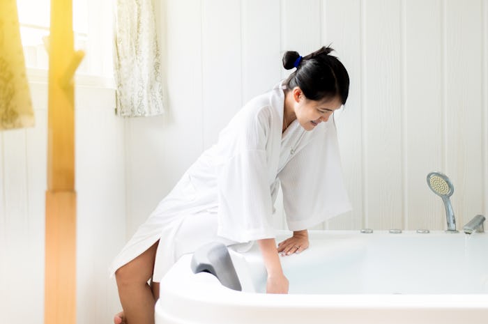 woman in white robe leaning against white tub feeling the water temperature