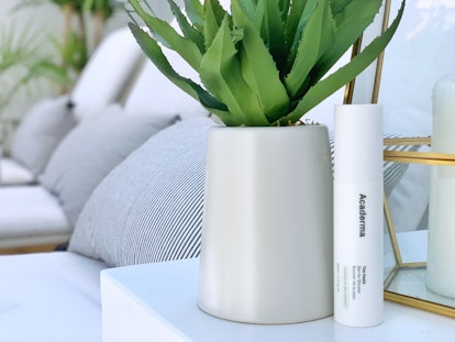 The Invisible Shield Defense Serum by Acaderma next to a vase with a plant on a white table