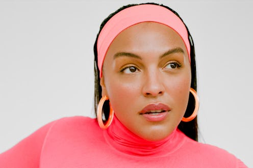 A brunette woman with a pink headband, pink turtleneck, and peach hoop earrings