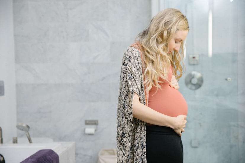 Woman in bathroom gazes down at pregnant bump.