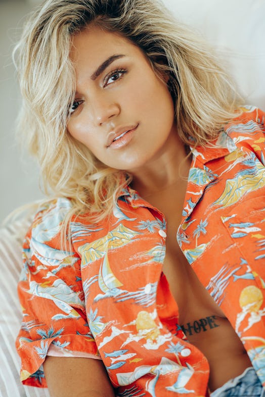 Karol G posing in an unbuttoned orange Hawaiian shirt showing off her tattoo