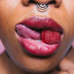 A woman with a nose ring and her mouth slightly opened revealing her tongue and a raspberry