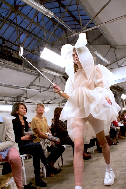The model is wearing a white frilled sheer dress and a supersized bow on her head