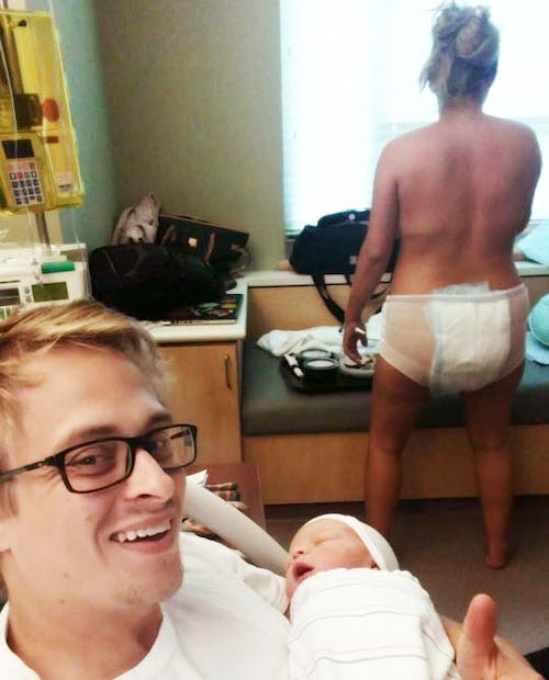 A man holding his newborn baby in the hospital while his wife stands in the background wearing mesh ...