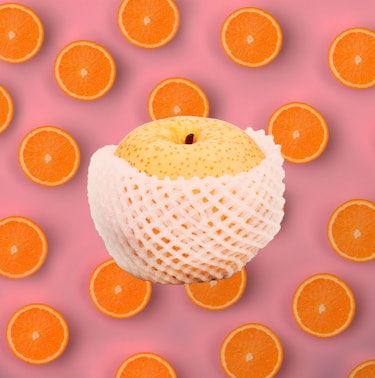 An apple in a mesh cover with a background of halved oranges