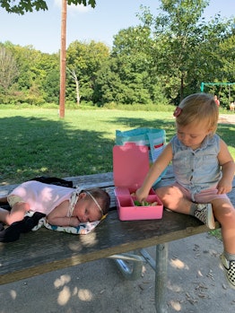 A baby lies on a picnic table next to a toddler